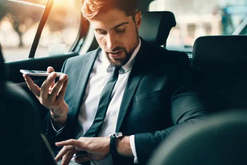 Businessman sitting in the back seat of a car, holding a smartphone for a voice message while checking the time on his wristwatch, dressed in a dark suit and tie. symbolizing Monterrey Black Car Service.
